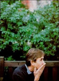 a man sitting on a wooden bench in front of trees and bushes with his hand to his mouth