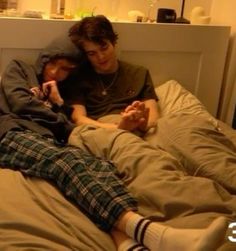 two young men laying in bed with one holding the other's hand and looking at his phone