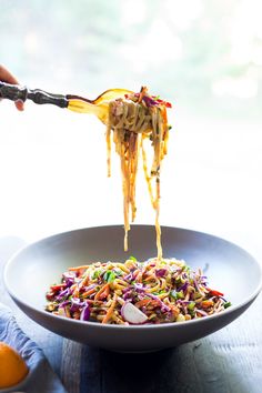 someone is lifting noodles from a bowl with carrots and an orange in the background