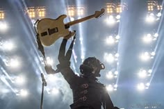 a man holding up a guitar in front of a crowd at a music concert with lights behind him