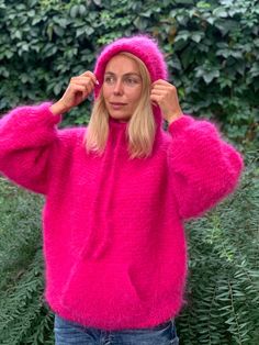 a woman wearing a bright pink fuzzy sweater and hoodie in front of some bushes