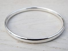 "A lovely solid sterling silver bangle, hand forged from 5.7mm X 3.0mm oval wire.  It can then be polished to a high shine finish or given a soft satin finish.   The shape, weight and smooth finish of this bangle makes it lovely and tactile.  It can be worn on its own or as part of a set of stacking bangles.  The average weight is 29g of silver. This bangle is hallmarked in the London Assay Office. The bangle is available as a circle or oval in the following sizes: SMALL   60mm  |  190mm or 7 1/2\" MEDIUM   65mm  |  204mm or 8\" LARGE   70mm  |  220m or 8 3/4\" Sizes shown are the inside diameter & circumference. Beautifully presented in a tinysilver faux suede pouch. Bangles help the planet!  For every bangle sold we plant a tree with JUST ONE Tree." Silver Oval Bangle With Polished Finish, Modern Silver Oval Bangle, Minimalist Oval Bangle As Gift, Minimalist Oval Bangle As A Gift, Minimalist Oval Bangle Gift, Mens Bangles, Stacking Bangles, Bangles Making, Sterling Silver Bangle