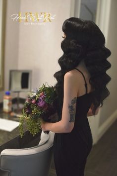 a woman in a black dress is holding a bouquet and looking at the bathroom mirror