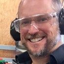 a man wearing headphones and glasses smiles at the camera in front of some electronics