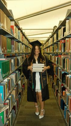 a woman in a graduation cap and gown is holding a book while standing in a library