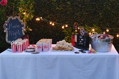 an outdoor movie party with popcorn, soda cans and snacks on a table in front of a hedge