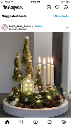 a small house surrounded by trees and lit candles on top of a white plate with pine cones