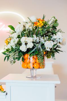 a vase filled with flowers and oranges on top of a white table next to a neon sign