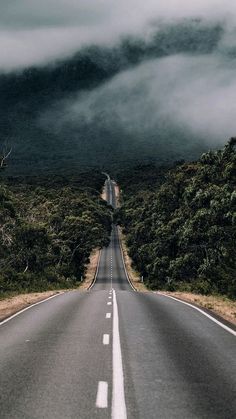 an empty road surrounded by trees and mountains with low clouds in the sky above it