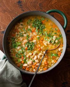 a pot filled with beans and carrots on top of a wooden table next to a spoon