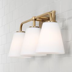 three light bathroom fixture with white glass shades on the bottom and gold finish, against a white tiled wall