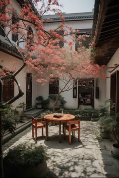 a table and two chairs under a tree with pink flowers in the middle of a courtyard