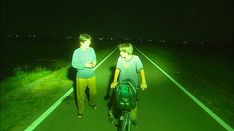 two men walking down the road at night with their bikes and backpacks in hand