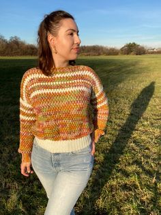 a woman is standing in the grass wearing a colorful sweater and jeans with her hands on her hips