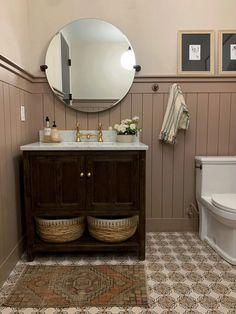 a white toilet sitting next to a bathroom sink under a round mirror on top of a wooden cabinet