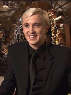 a man with blonde hair wearing a black suit and smiling at the camera while sitting down