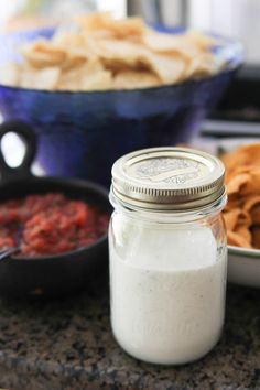 a glass jar filled with white liquid next to bowls of chips and salsa on a counter