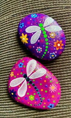 two painted rocks with flowers and dragonflies on them, sitting on a brown surface