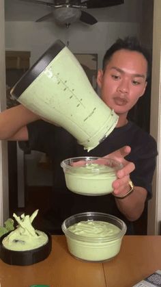 a man pouring green liquid into a bowl on top of a wooden table in front of a fan