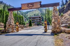 an entrance to a resort surrounded by trees and mountains in the distance is a road with stone pillars leading into it