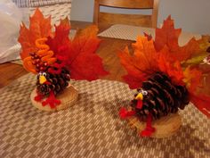 two turkeys made out of pine cones and autumn leaves on a table with a chair in the background