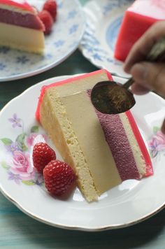 a piece of cake on a plate with raspberries in the foreground and someone cutting into it