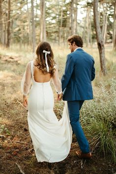 a bride and groom walking through the woods holding hands