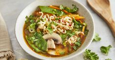 a white bowl filled with noodles and vegetables next to a wooden spoon on top of a table