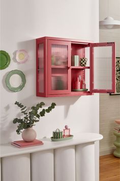 a red cabinet sitting on top of a white counter next to a plant and plates