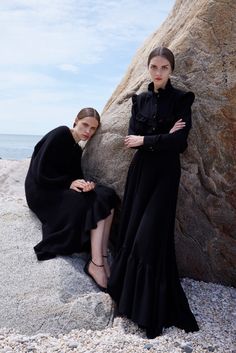 two women in black dresses sitting on rocks near the ocean