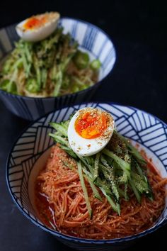 two bowls filled with noodles and vegetables on top of a black countertop next to an egg