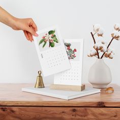 a person holding a calendar next to a small white vase with cotton flowers in it