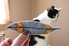 a cat is sitting in the background while someone holds a toy fish that looks like a feather