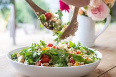 a person is sprinkling vegetables into a salad in a white bowl on a wooden table