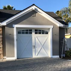 a garage with two white doors and brick walkway leading up to the front door, next to a palm tree