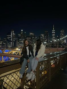 two women sitting on a railing in front of a city skyline at night with the lights turned on