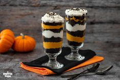 two glasses filled with dessert sitting on top of a black and orange napkin next to an orange pumpkin