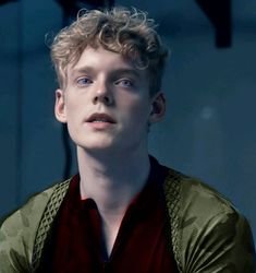 a young man with curly hair wearing a red shirt and green cardigan jacket looking at the camera