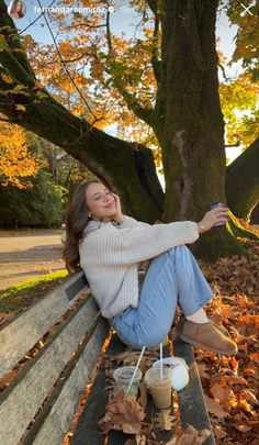 a woman is sitting on a bench in the fall leaves and holding a cup of coffee