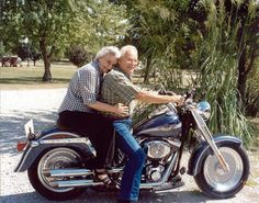 an older couple riding on the back of a motorcycle in front of trees and grass