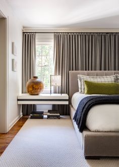 a bed sitting next to a window with curtains on the windowsill and a table in front of it