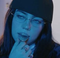 a woman wearing glasses and a beanie is holding her finger to her mouth while looking at the camera