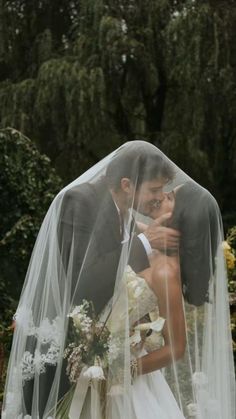 a bride and groom kissing under a veil in front of some trees on their wedding day