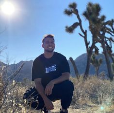 a man squatting down in the desert with trees and mountains behind him on a sunny day