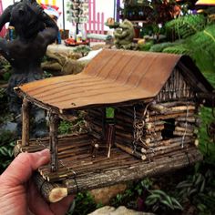 a hand holding a miniature house made out of wood and logs in front of plants