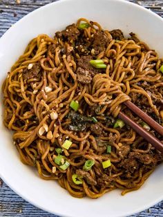 a white bowl filled with noodles and meat on top of a wooden table next to chopsticks