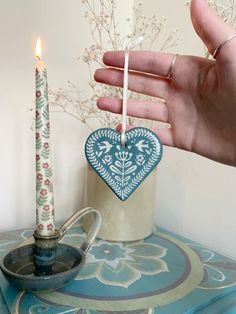 a hand holding a heart shaped ornament next to a tea cup and candle