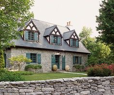 a stone house with green shutters on the front and side windows, surrounded by greenery