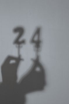 the shadow of a person's hand holding a clock with numbers on it in front of a white wall