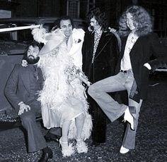 four people posing for a photo in front of a car with one person wearing a feathered costume